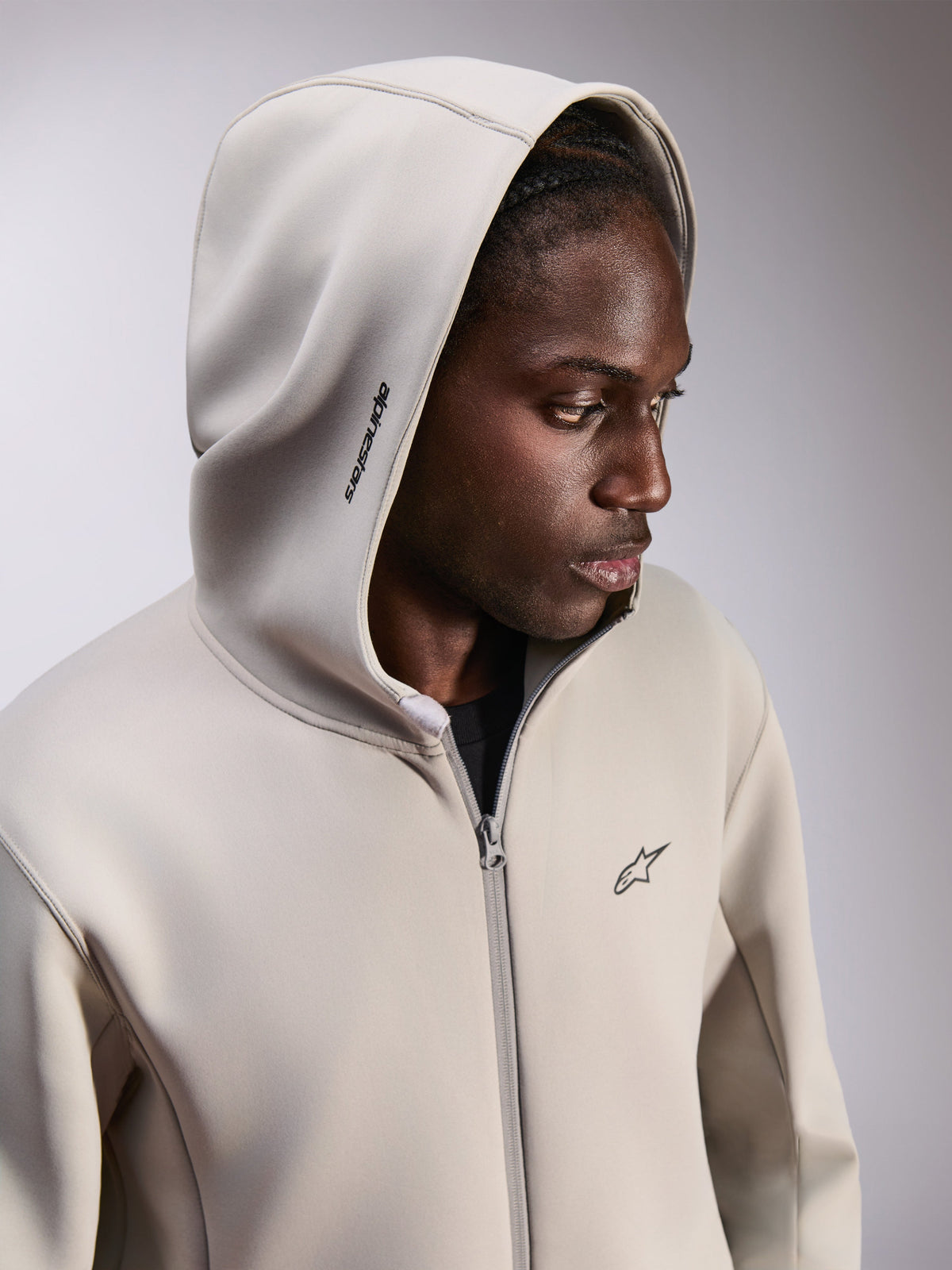 Alpinestars Cypher Hoodie, felpa in pile con zip, grigio cemento, indossata con il cappuccio alzato, con logo Alpinestars sul cappuccio e piccolo logo Astars sul petto, streetwear casual ispirato al motorsport