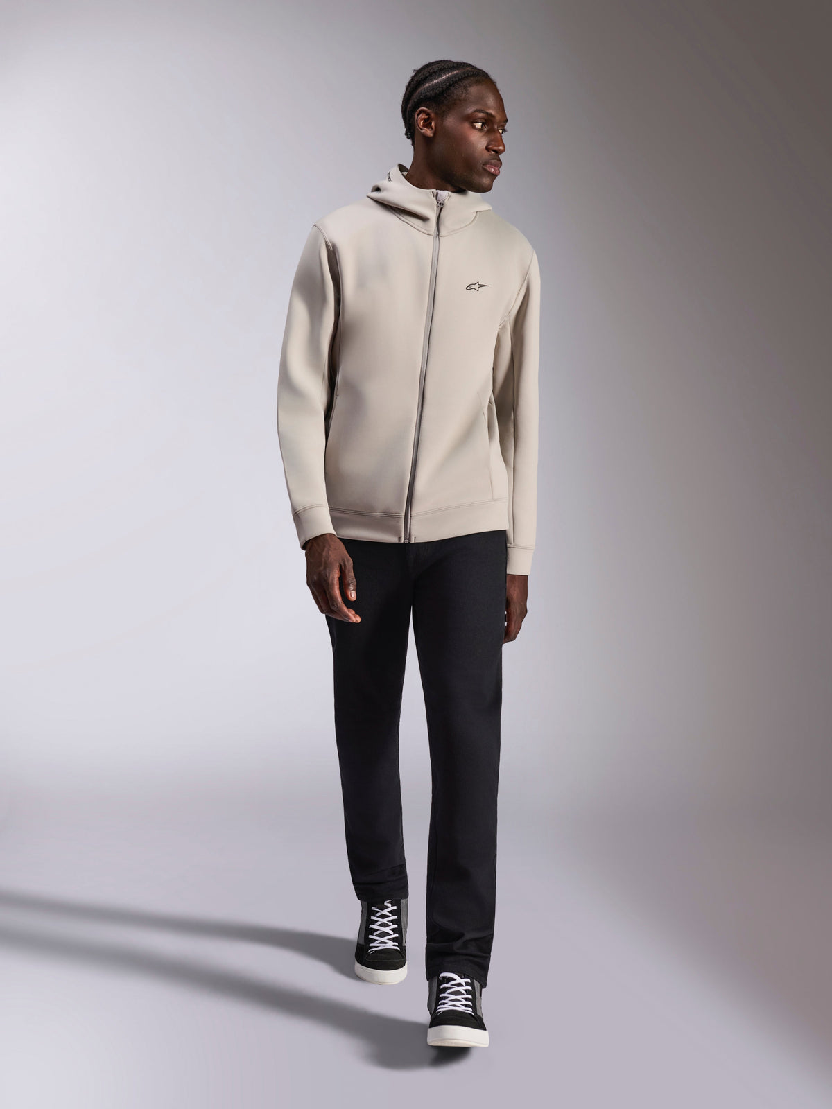 Alpinestars Cypher Hoodie, felpa con cappuccio in pile double face con zip integrale, grigio cemento, indossata da un modello con pantaloni neri e sneakers bianche e nere, dotata di cappuccio a collo alto foderato in rete e piccolo logo Astars sul petto, streetwear casual moderno