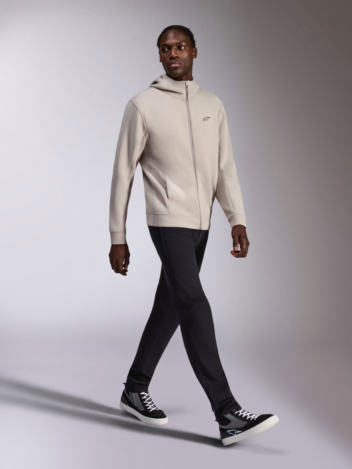 Felpa con cappuccio Alpinestars Cypher, pile con zip integrale grigio cemento, cappuccio a collo alto, piccolo logo Astars sul petto, indossata da un modello che cammina, abbinata a pantaloni neri e sneakers nere e grigie, abbigliamento sportivo casual ispirato al motorsport.