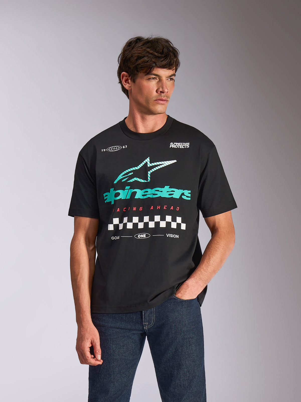 T-shirt Alpinestars Aplomb Oversized CSF, maglietta a maniche corte, nera, con un grande logo combinato Astars verde acqua e bianco e grafica a scacchi, indossata da un modello uomo con jeans blu scuro, stile streetwear casual ispirato al motorsport