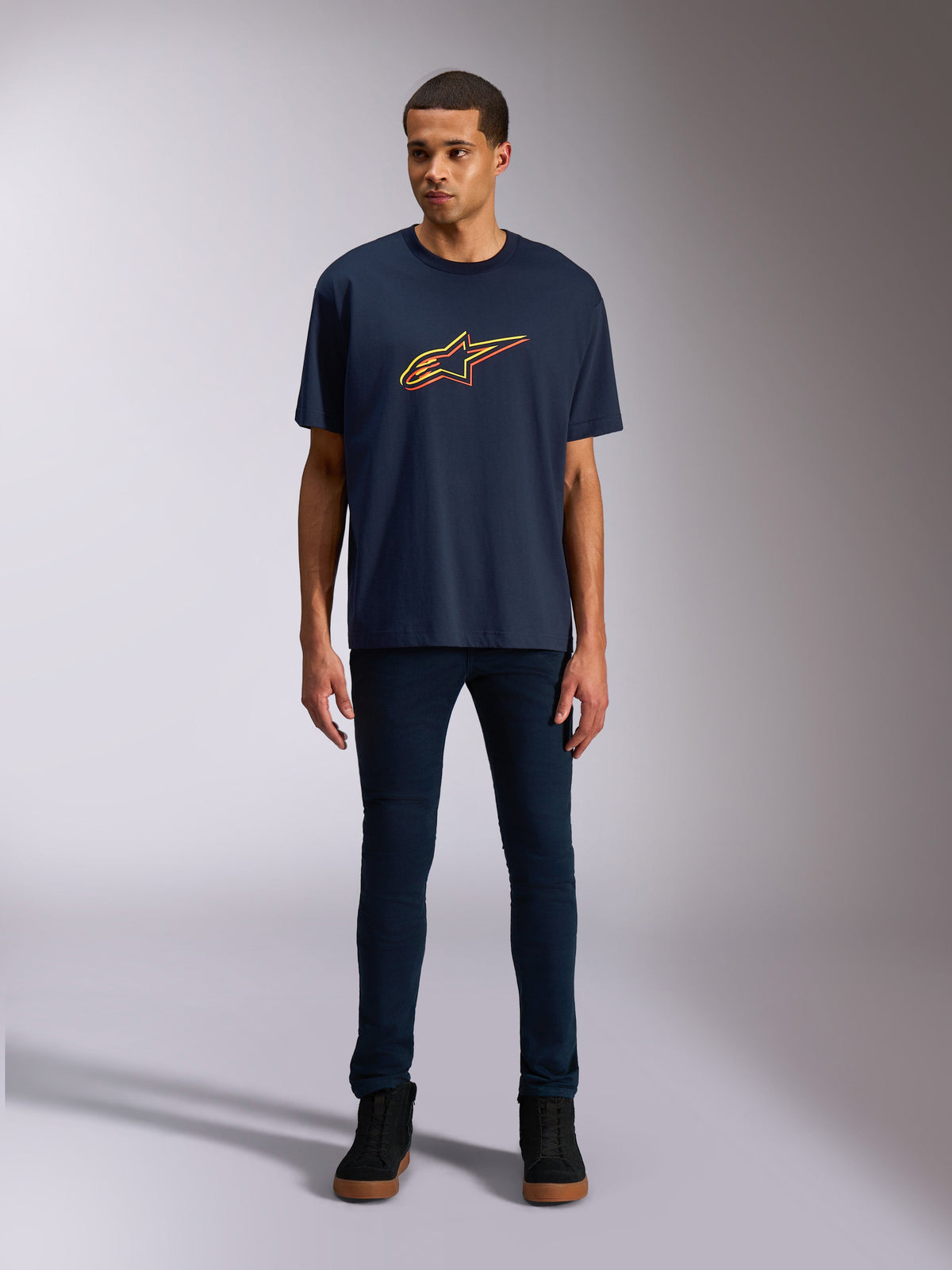 T-shirt Alpinestars Alter Oversized CSF, maglietta a maniche corte, blu navy, con logo Alpinestars a stella frontale con colore sfalsato giallo e arancione, indossata da un modello con denim scuro e sneakers nere, abbigliamento sportivo casual lifestyle.
