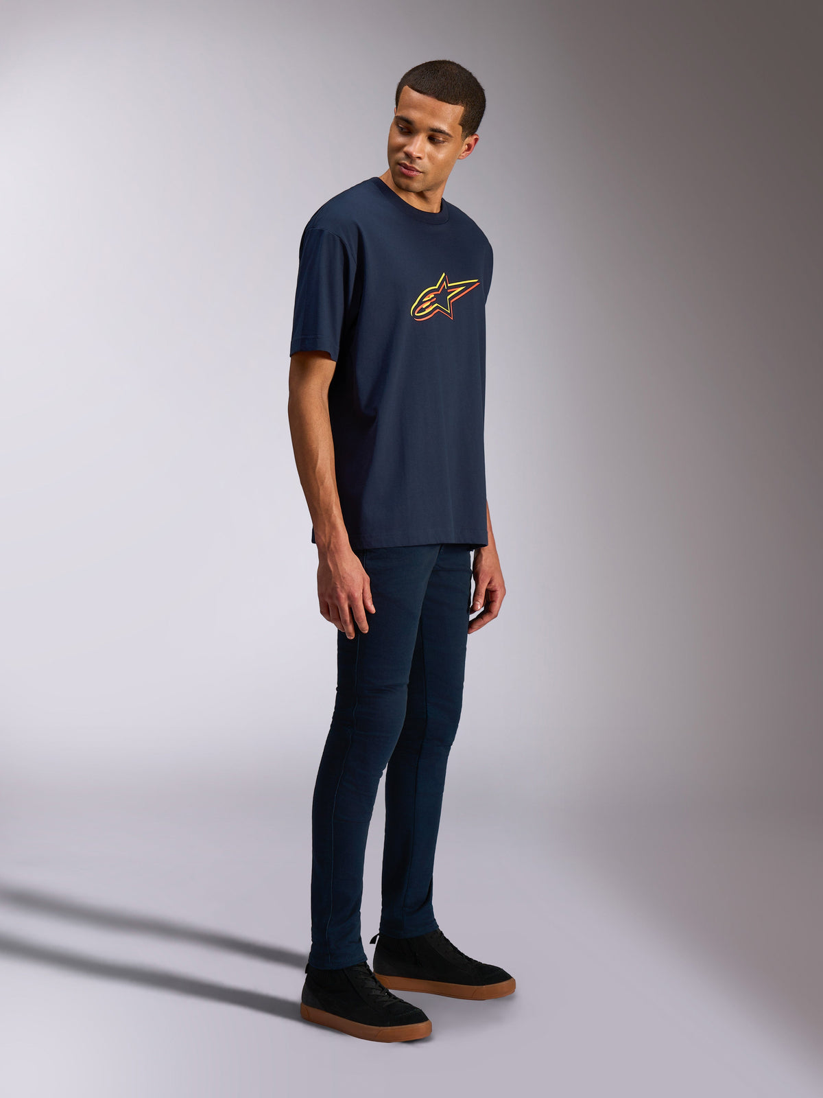 Alpinestars Alter Oversized CSF Tee, t-shirt a maniche corte, blu navy, logo Astars giallo e arancione, indossata da un modello con jeans scuri e sneakers nere, streetwear casual ispirato al motorsport