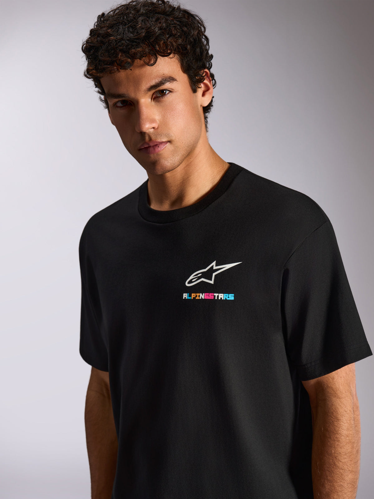 Un uomo indossa la T-shirt Alpinestars Wild Oversized CSF, maglietta nera a maniche corte con logo Astars bianco e branding multicolore vivace sul petto, streetwear casual motorsport