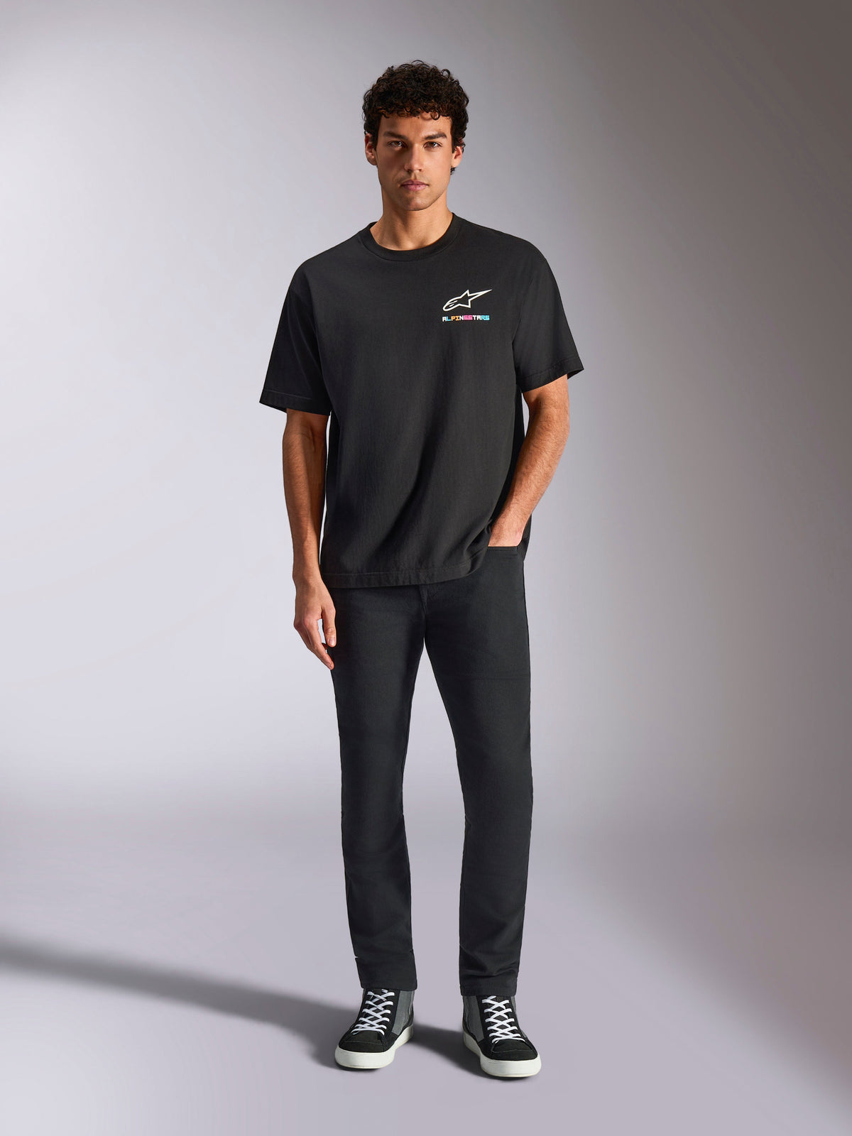 Alpinestars Wild Oversized CSF Tee, t-shirt a maniche corte, nero, indossata da un modello con pantaloni neri e sneakers, con logo Astars bianco e branding multicolore vivace, streetwear casual ispirato al motorsport
