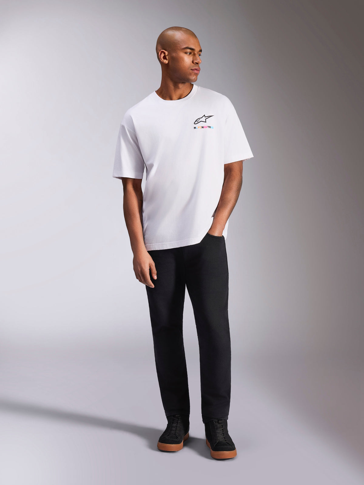 Alpinestars Wild Oversized CSF Tee, t-shirt a maniche corte, bianca, logo Astars multicolore vivace sul petto, indossata con pantaloni neri e sneakers, abbigliamento lifestyle casual motorsport