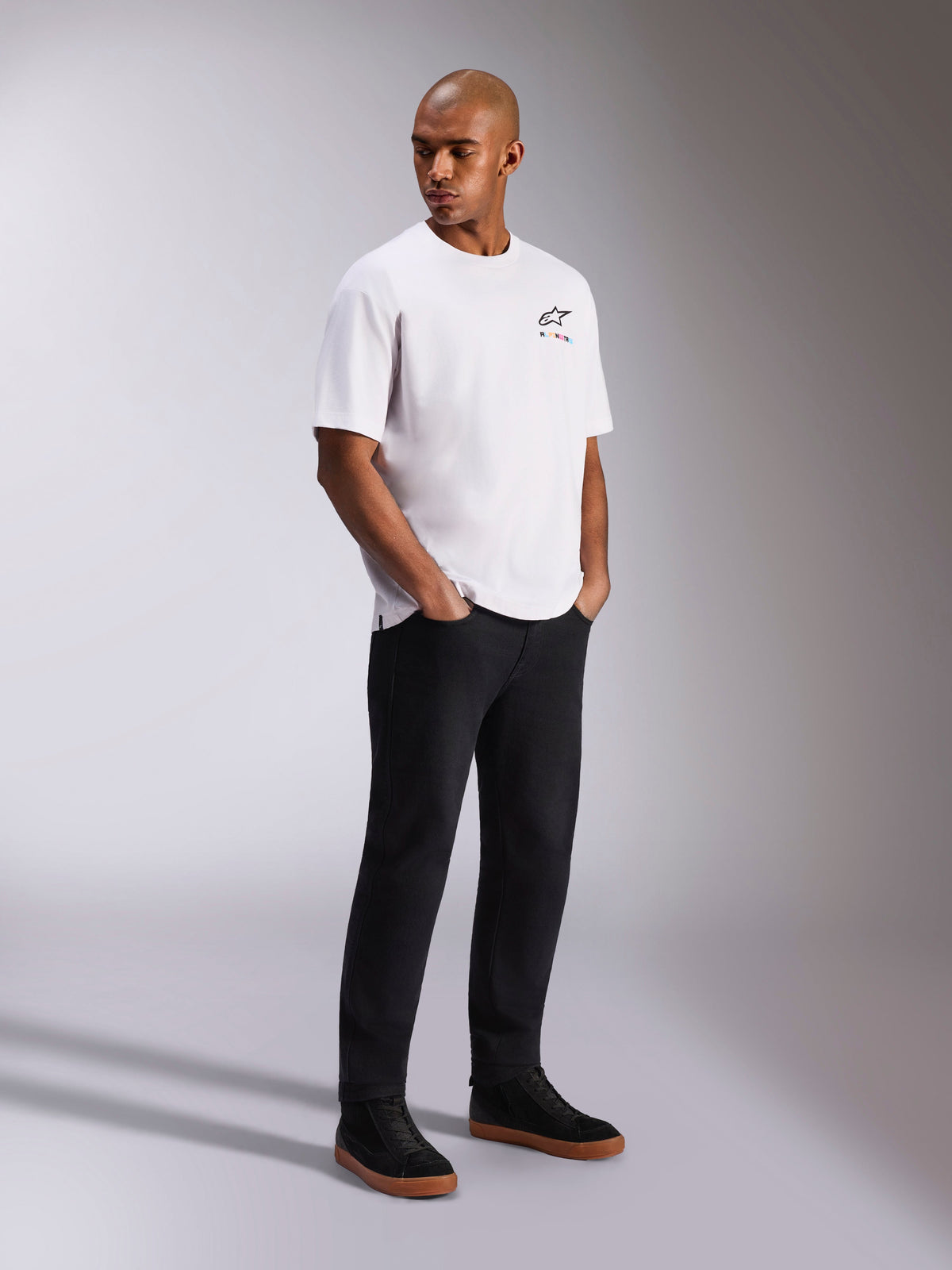 Alpinestars Wild Oversized CSF Tee, maglietta a maniche corte, bianca, con un piccolo logo Astars multicolore sul petto, indossata da un modello con pantaloni neri e sneaker nere, streetwear casual ispirato al motorsport