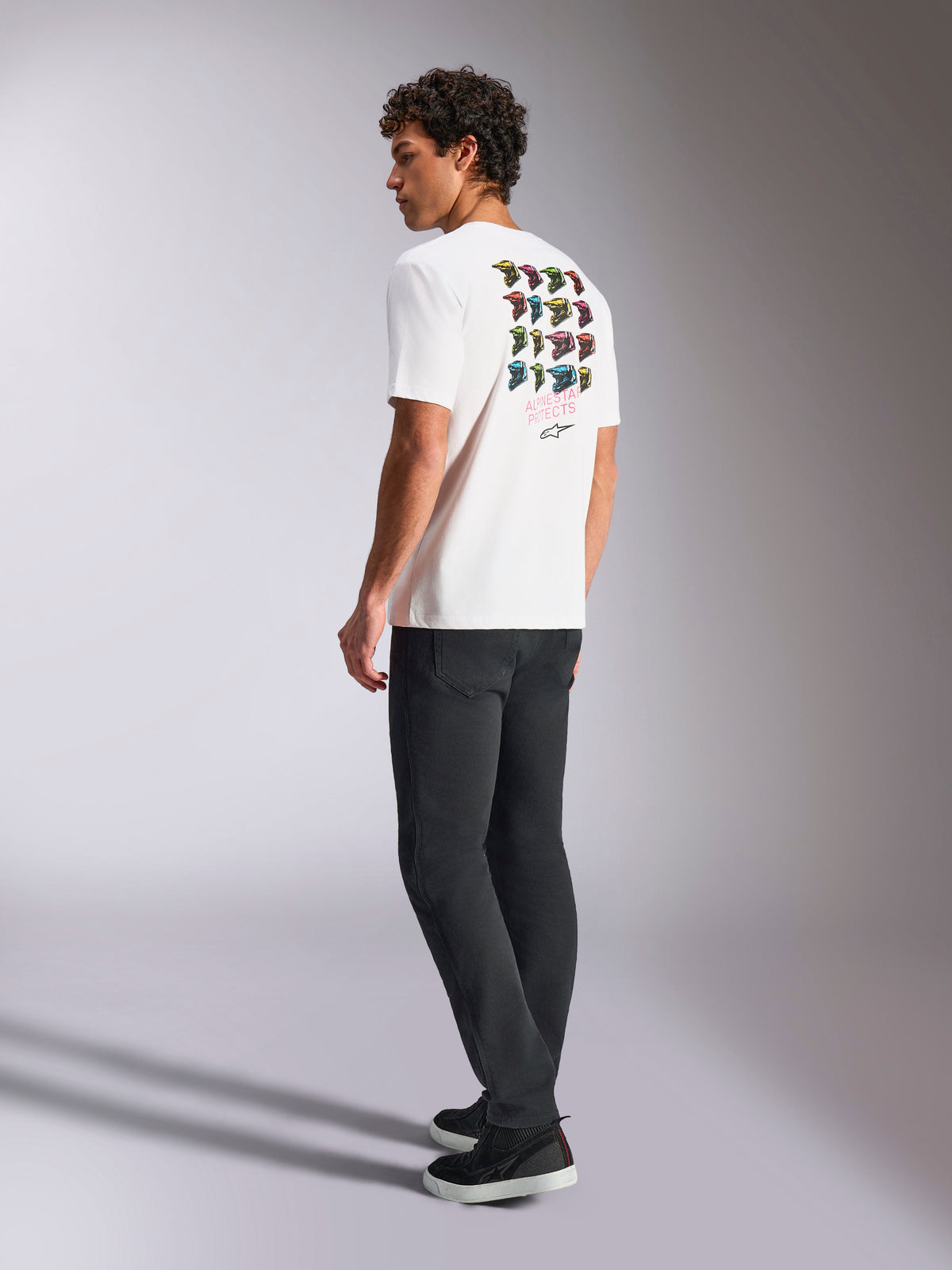 Maglietta Alpinestars Pop Helmet CSF, t-shirt a maniche corte, bianca, vista posteriore su modello con grafica pop colorata dei caschi M10, abbinata a pantaloni scuri e sneakers nere, streetwear casual ispirato al motorsport