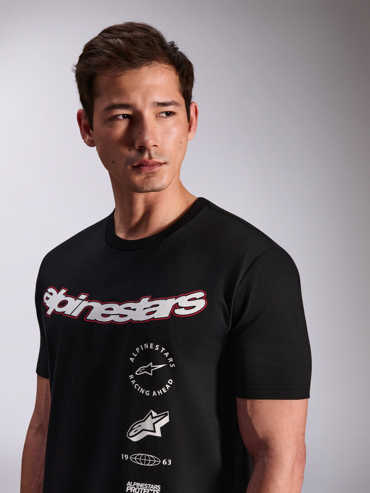Alpinestars Abus CSF Tee, T-shirt a maniche corte, nera, indossata da un modello, con logo Alpinestars bianco con bordo rosso e loghi grafici multipli impilati sul petto, streetwear casual motorsport