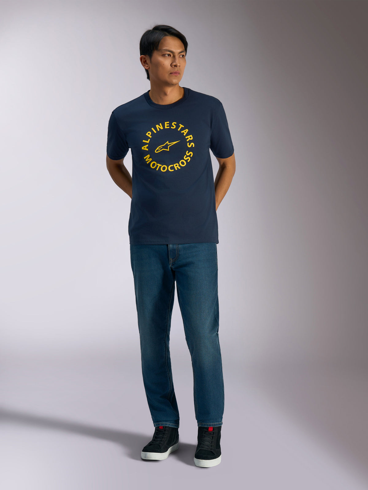 Alpinestars MX CSF Tee, t-shirt a maniche corte, blu navy, con grafica circolare Alpinestars Motocross gialla, indossata da un modello con jeans blu e sneakers nere, streetwear casual motorsport