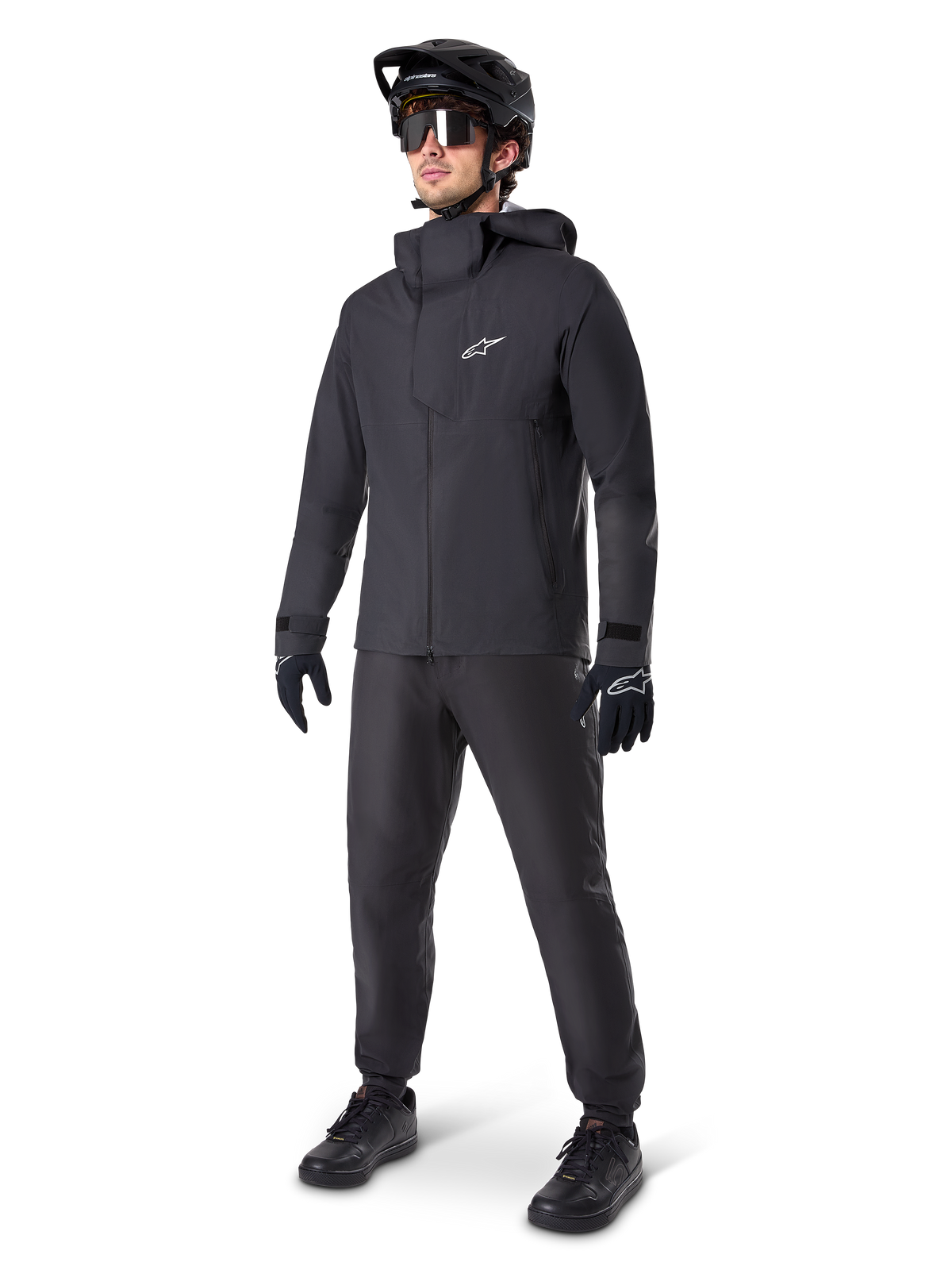 Giacca impermeabile Alpinestars A-Dura Elite in nero, indossata da un pilota uomo con pantaloni da MTB, guanti e casco neri coordinati, design tecnico impermeabile, logo Astars bianco sul petto e collo alto protettivo.