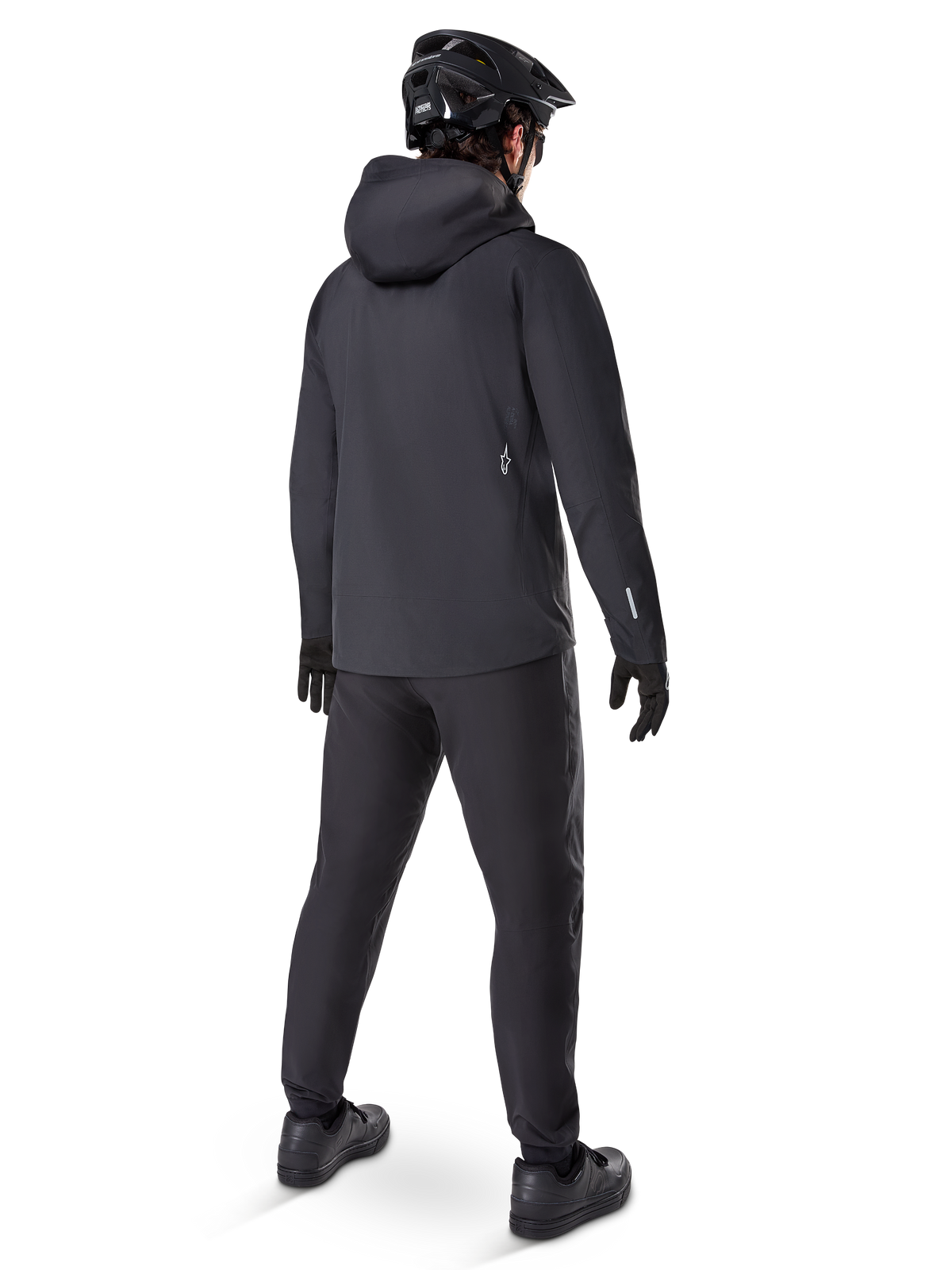Giacca impermeabile Alpinestars A-Dura Elite, Nero, vista posteriore con cappuccio sopra il casco e orlo posteriore più lungo, abbinata a pantaloni MTB neri coordinati, guanti e casco per una protezione completa sui sentieri in ogni condizione atmosferica.