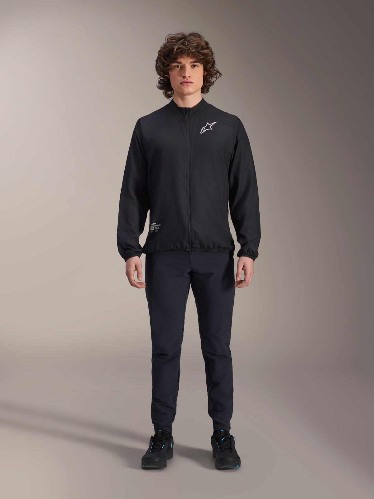 Modello che indossa la giacca frangivento Alpinestars A-Aria in nero, con cerniera frontale integrale, collo alto e logo Astars bianco sul petto, abbinata a pantaloni tecnici neri e scarpe nere, abbigliamento tecnico da ciclismo per mountain bike e uso outdoor.