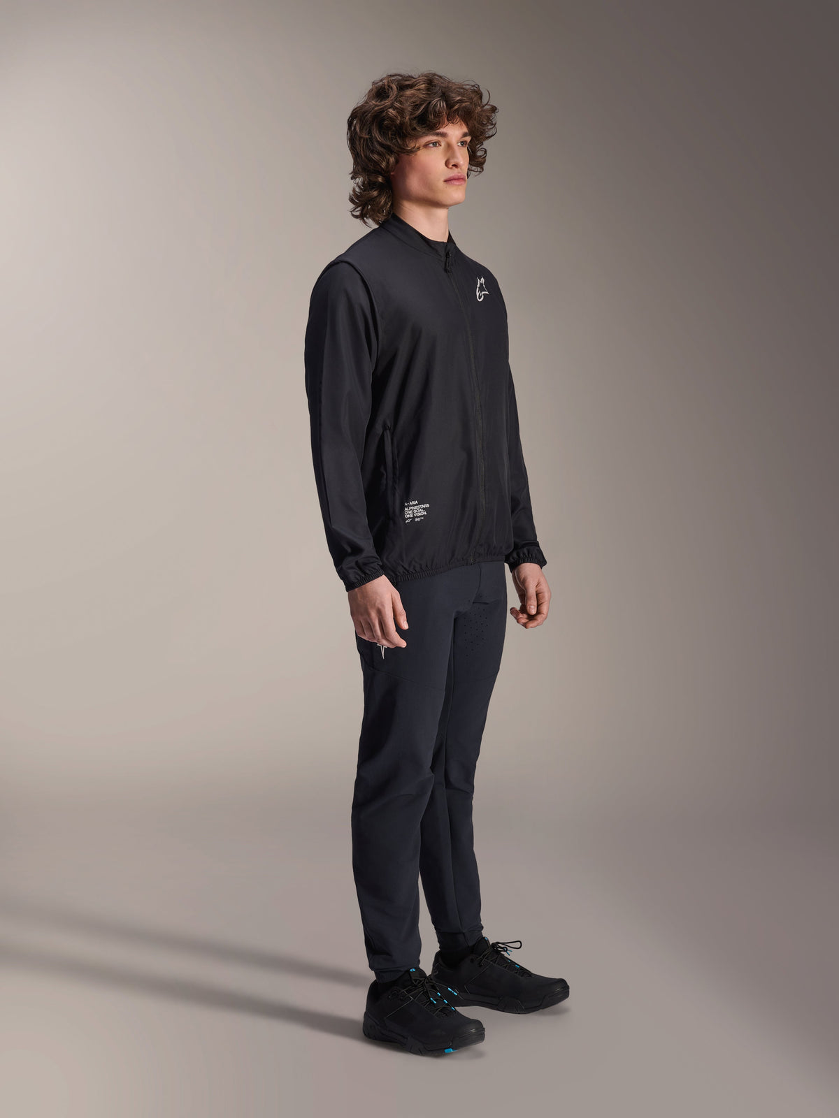 Giacca frangivento Alpinestars A-Aria, nera, giacca da ciclismo leggera e resistente al vento indossata da un modello con capelli ricci, con logo Astars bianco sul petto e marchio in basso a destra, abbinata a pantaloni e scarpe neri, vestibilità performante per mountain bike e gravel