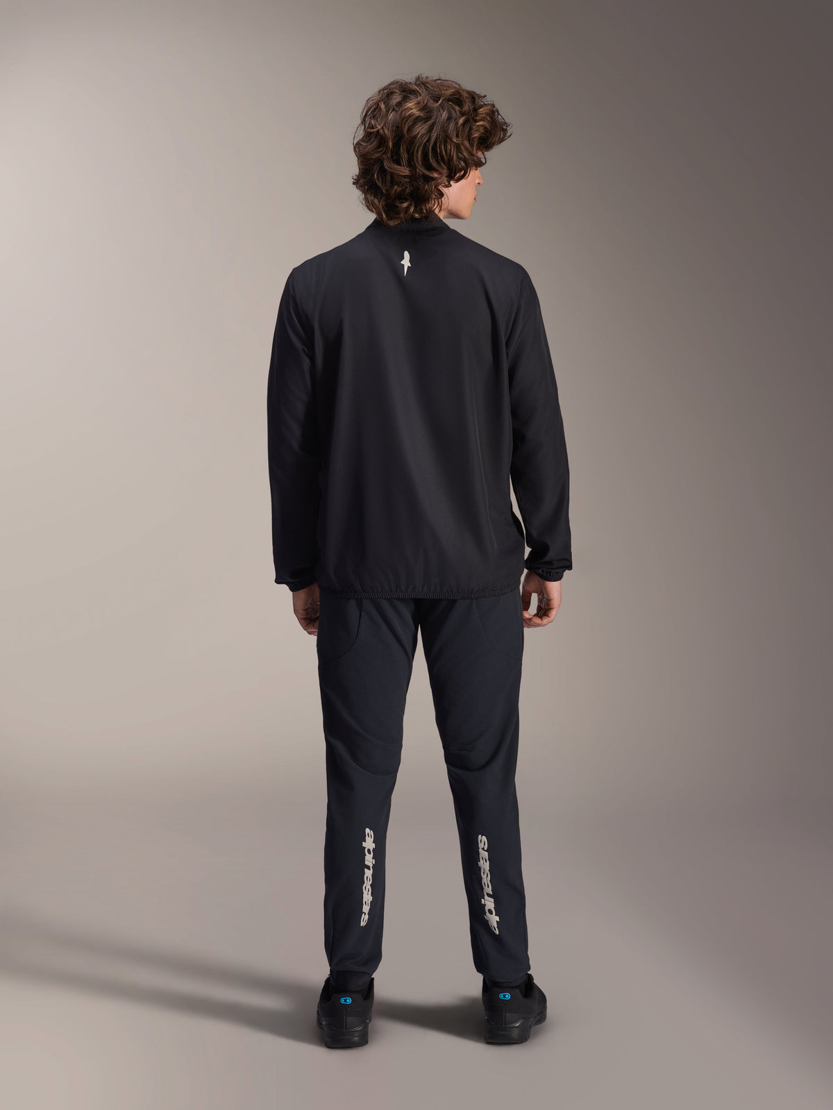 Giacca frangivento Alpinestars A-Aria, nero, vista posteriore, giacca da ciclismo leggera e resistente al vento con un piccolo logo bianco sulla parte superiore della schiena, indossata con pantaloni neri coordinati con marchio Alpinestars verticale sui polpacci, abbigliamento tecnico da mountain bike