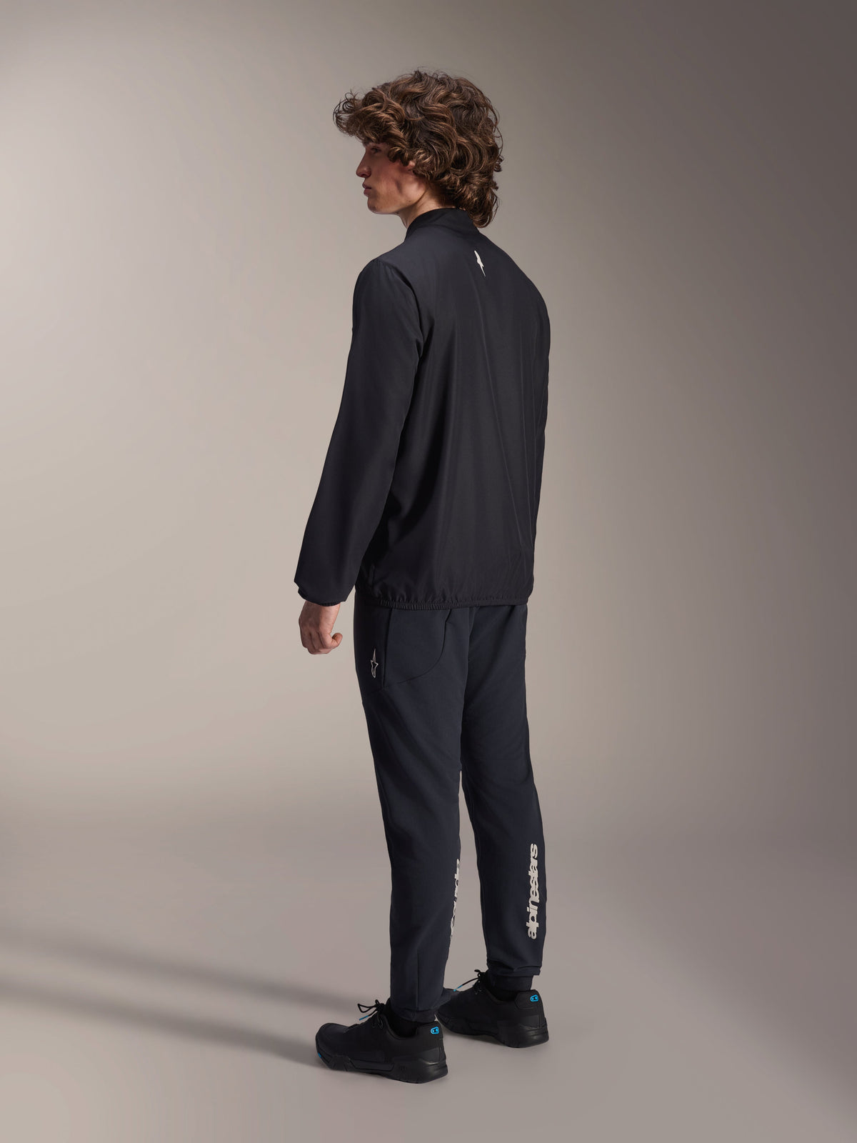 Giacca frangivento Alpinestars A-Aria, nera, giacca da ciclismo leggera e resistente al vento, vista posteriore di tre quarti su modello, con un piccolo logo bianco sul retro del collo e bordo elasticizzato, abbinata a pantaloni e scarpe neri coordinati per un look orientato alle prestazioni.