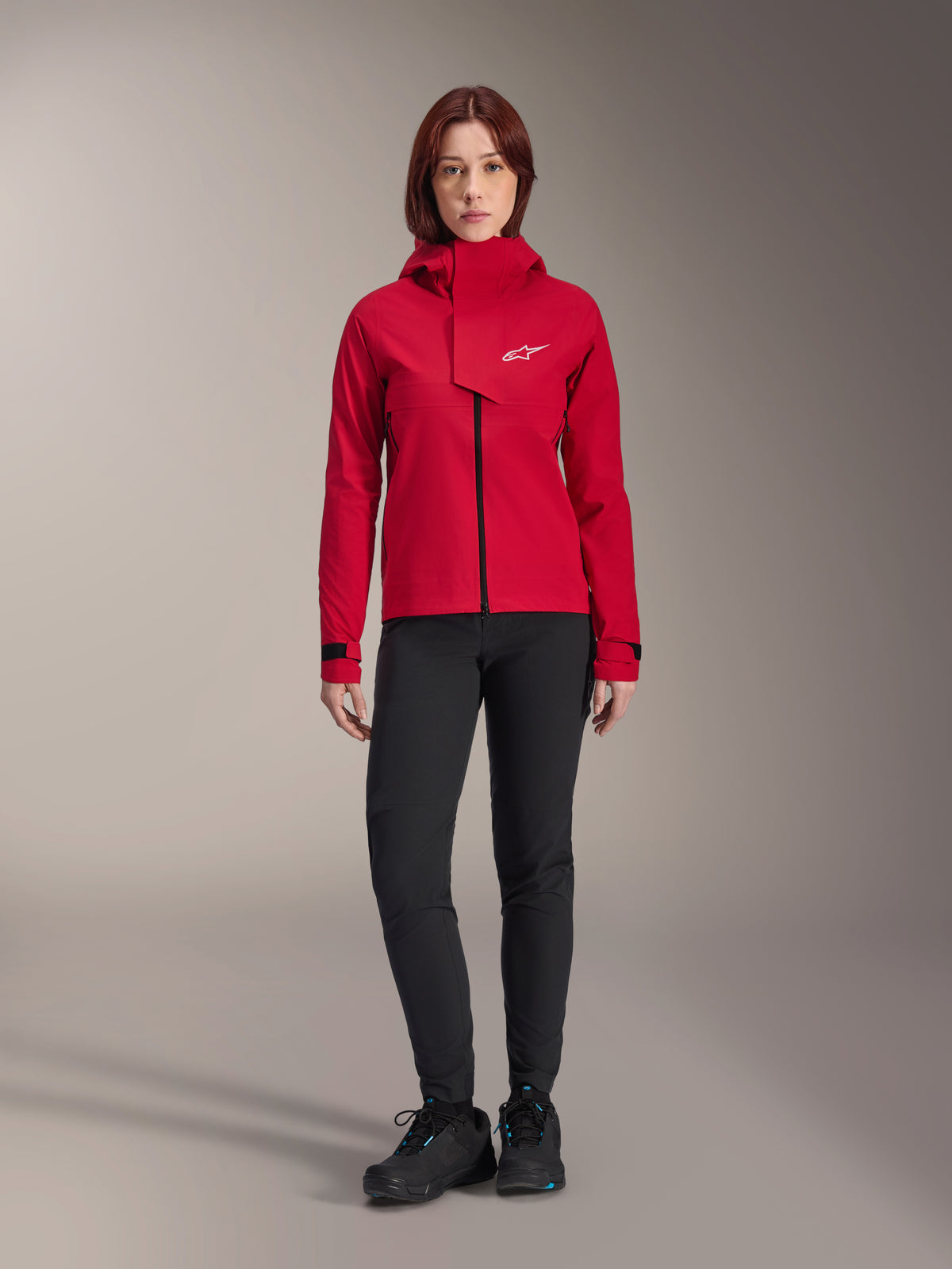 Giacca impermeabile Alpinestars Stella A-Dura Elite, giacca da MTB per donna, Rosso Fluo, indossata con pantaloni e scarpe neri, con cappuccio a collo alto e logo Alpinestars bianco, abbigliamento tecnico da mountain bike
