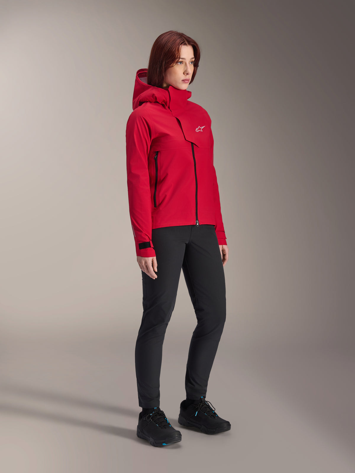 Giacca impermeabile Alpinestars Stella A-Dura Elite da donna, Rosso Fluo, giacca antipioggia con cappuccio per mountain bike con logo Astars bianco sulla patta pettorale, indossata con pantaloni neri e scarpe da mountain bike nere, abbigliamento tecnico ad alte prestazioni per trail riding
