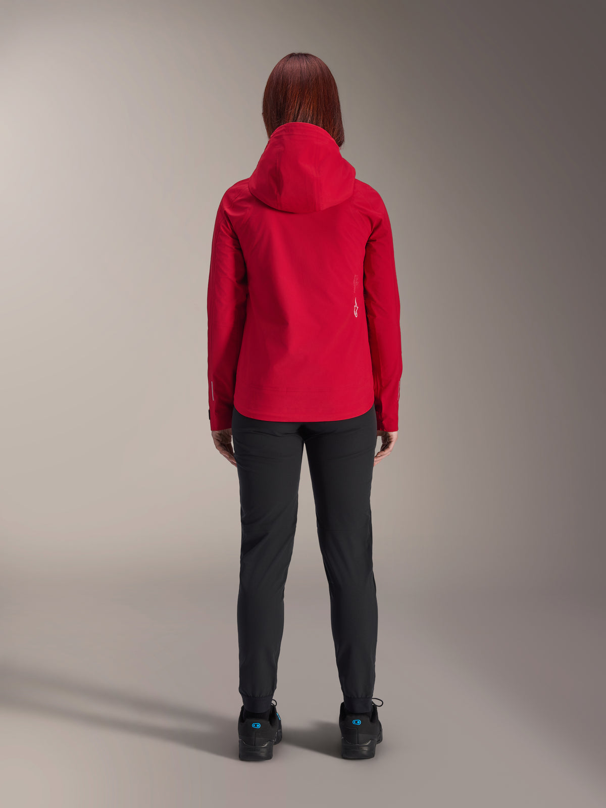 Vista posteriore di una donna che indossa la giacca impermeabile Alpinestars Stella A-Dura Elite in Red Fluo, abbinata a pantaloni e scarpe neri, dotata di un cappuccio protettivo e orlo posteriore allungato per la mountain bike.