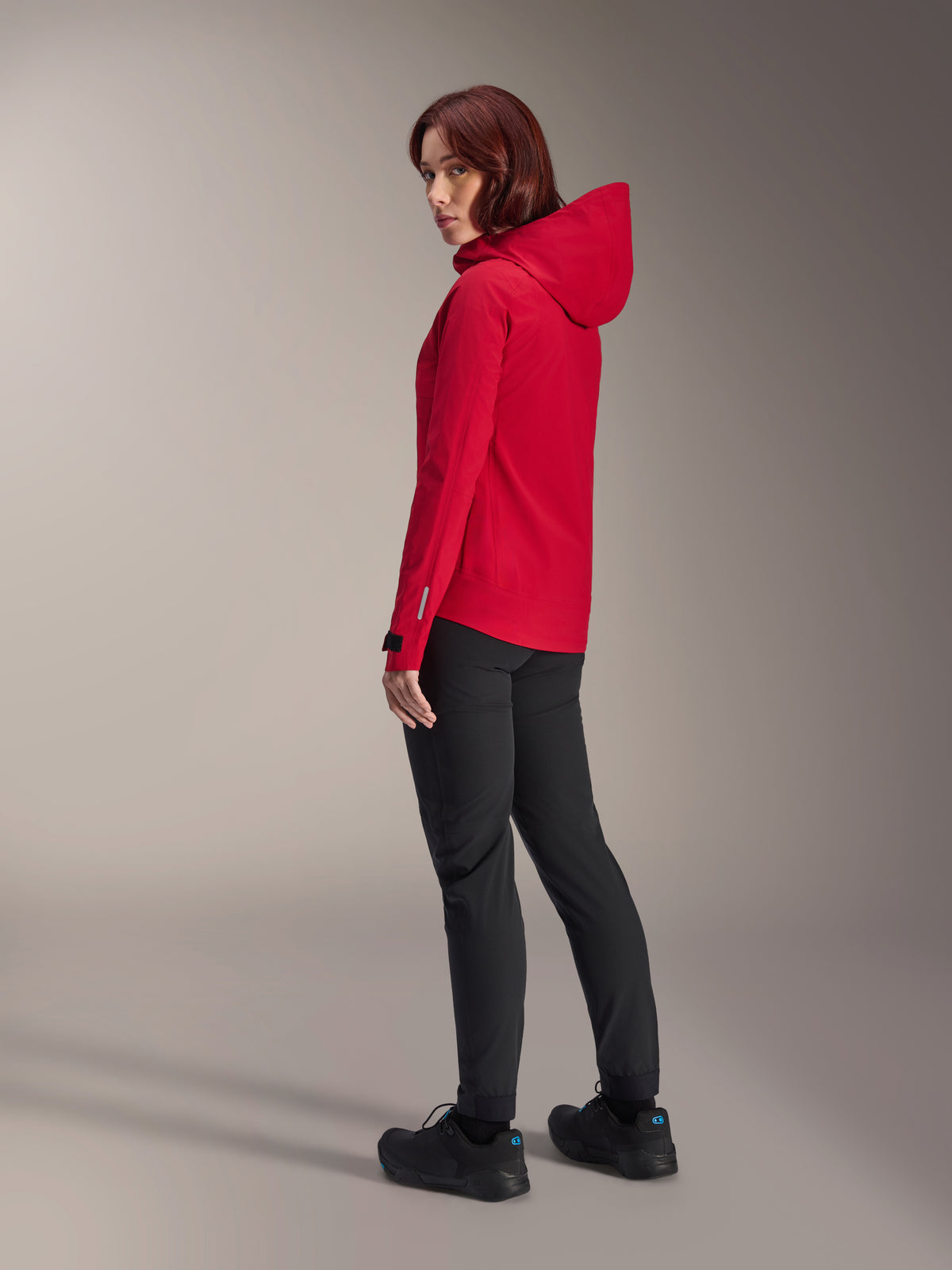 Donna che indossa la giacca impermeabile Alpinestars Stella A-Dura Elite in Red Fluo, abbinata a pantaloni tecnici da riding neri e scarpe nere, vista posteriore di tre quarti che mostra il cappuccio regolabile e l'orlo posteriore allungato, abbigliamento tecnico per mountain bike