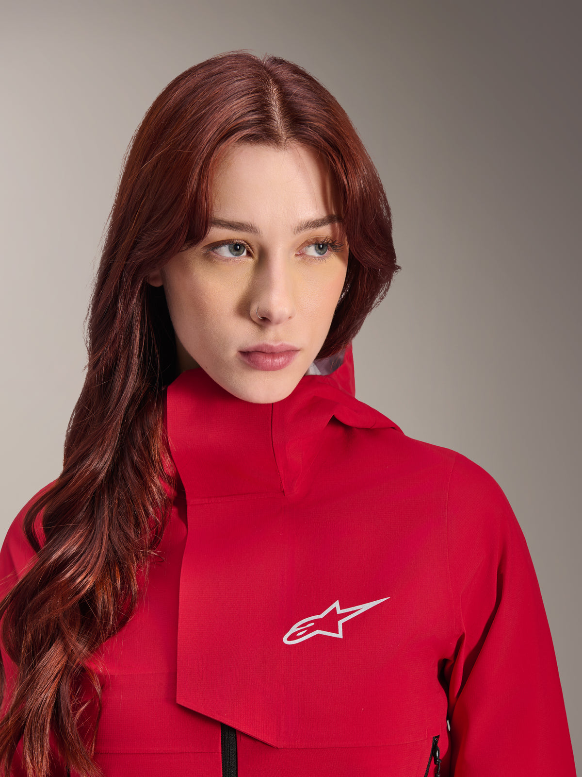 Alpinestars Stella A-Dura Elite Waterproof Jacket, rosso fluo, indossata da una donna con lunghi capelli rossi, giacca tecnica impermeabile a 3 strati con collo alto e logo bianco, equipaggiamento tecnico per outdoor e mountain bike