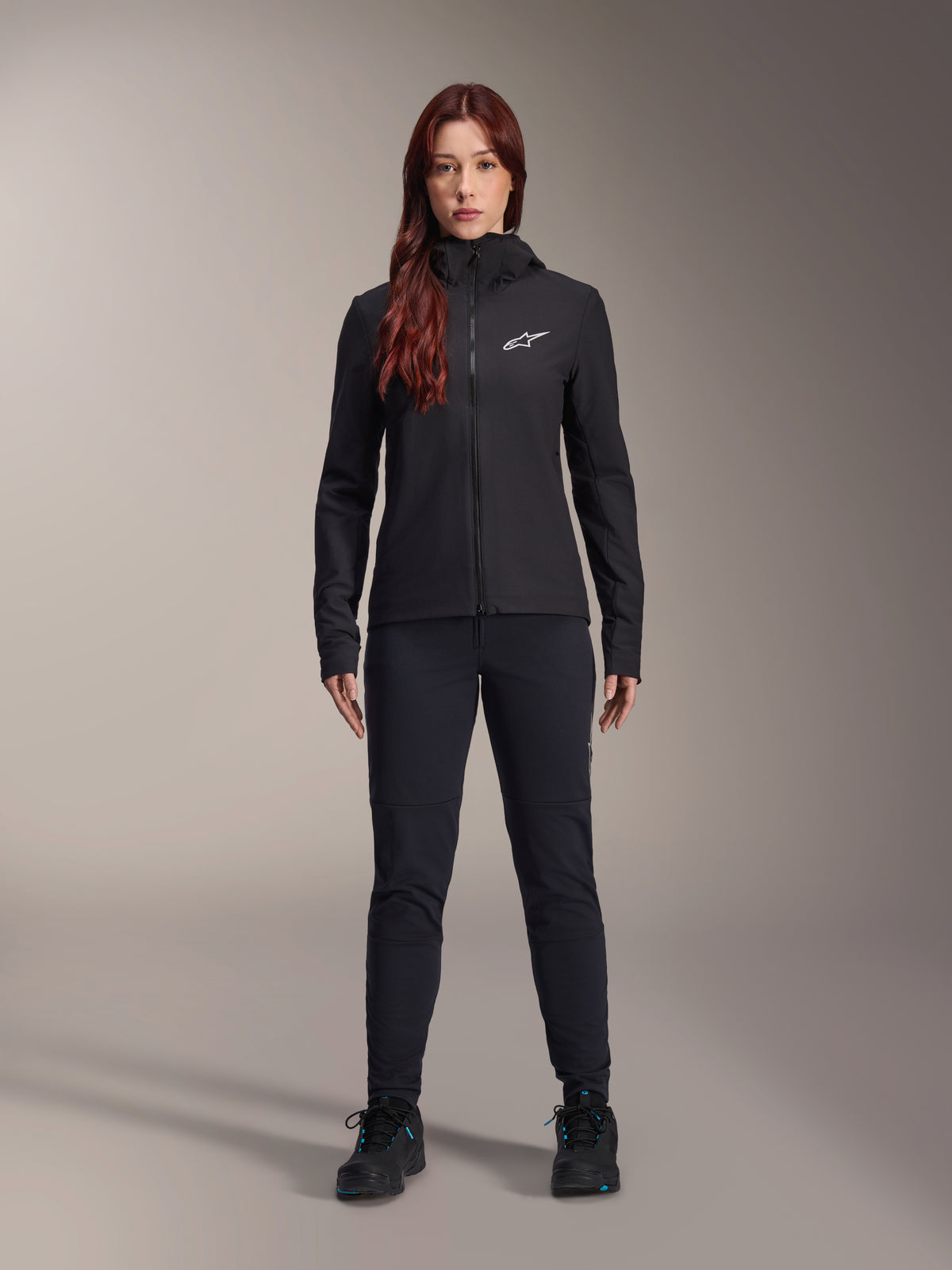 Donna che indossa la giacca termica Alpinestars Stella A-Dura, giacca da ciclismo leggera e isolata nera con cappuccio e logo Astars bianco sul petto, abbinata a pantaloni neri e scarpe sportive
