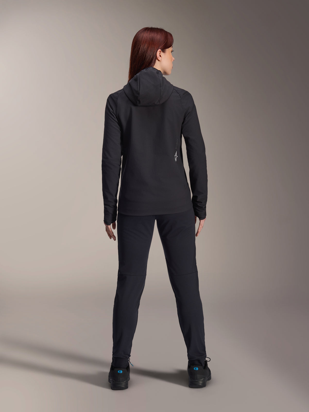 Donna con Giacca Termica Alpinestars Stella A-Dura, vista posteriore, giacca da ciclismo termica nera con cappuccio, pantaloni e scarpe neri coordinati, piccolo logo bianco sulla parte inferiore della schiena