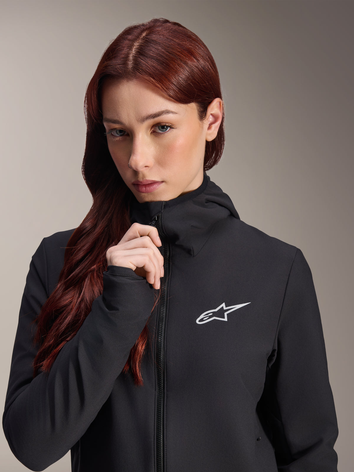 Giacca termica Alpinestars Stella A-Dura, giacca da ciclismo nera da donna, indossata da una modella con lunghi capelli castano-rossicci, logo Alpinestars bianco sul petto, design con cappuccio e zip integrale, abbigliamento da mountain bike