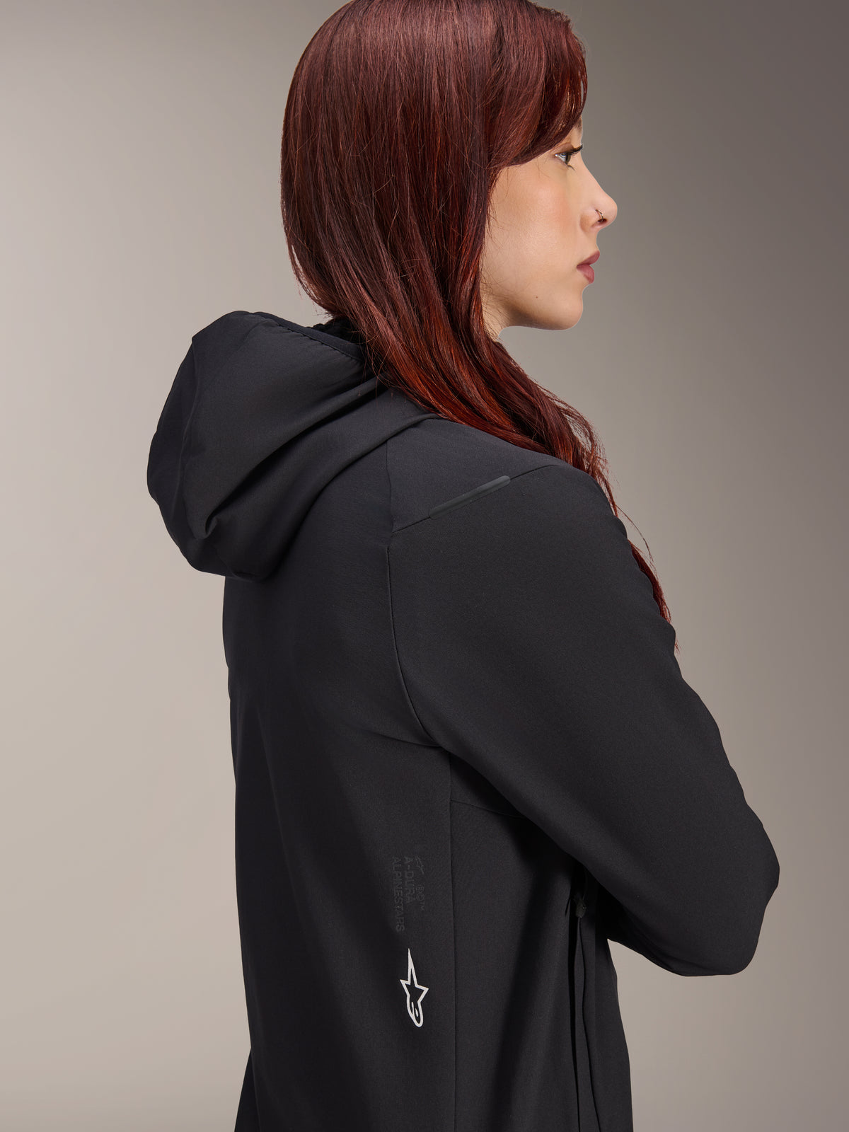 Giacca Termica Alpinestars Stella A-Dura, giacca da ciclismo da donna, nero, profilo laterale di una donna con i capelli rossi che indossa la giacca, dotata di cappuccio integrato e logo a stella Alpinestars bianco sul lato, abbigliamento termico isolante leggero per MTB