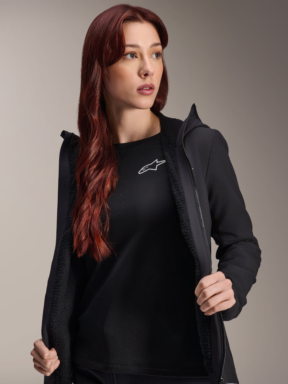 Giacca Termica Alpinestars Stella A-Dura, giacca da ciclismo nera da donna, mostrata aperta con fodera in pile testurizzato e zip frontale, indossata sopra una t-shirt nera con logo Alpinestars, leggera e isolata per la guida con il freddo