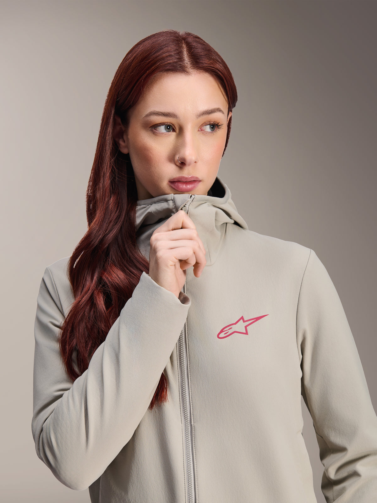Giacca termica da donna Alpinestars Stella A-Dura, grigio titanio con logo Alpinestars a stella rosa, modella con lunghi capelli rossi che indossa e chiude la zip della giacca con cappuccio, abbigliamento da ciclismo leggero e isolante per il freddo