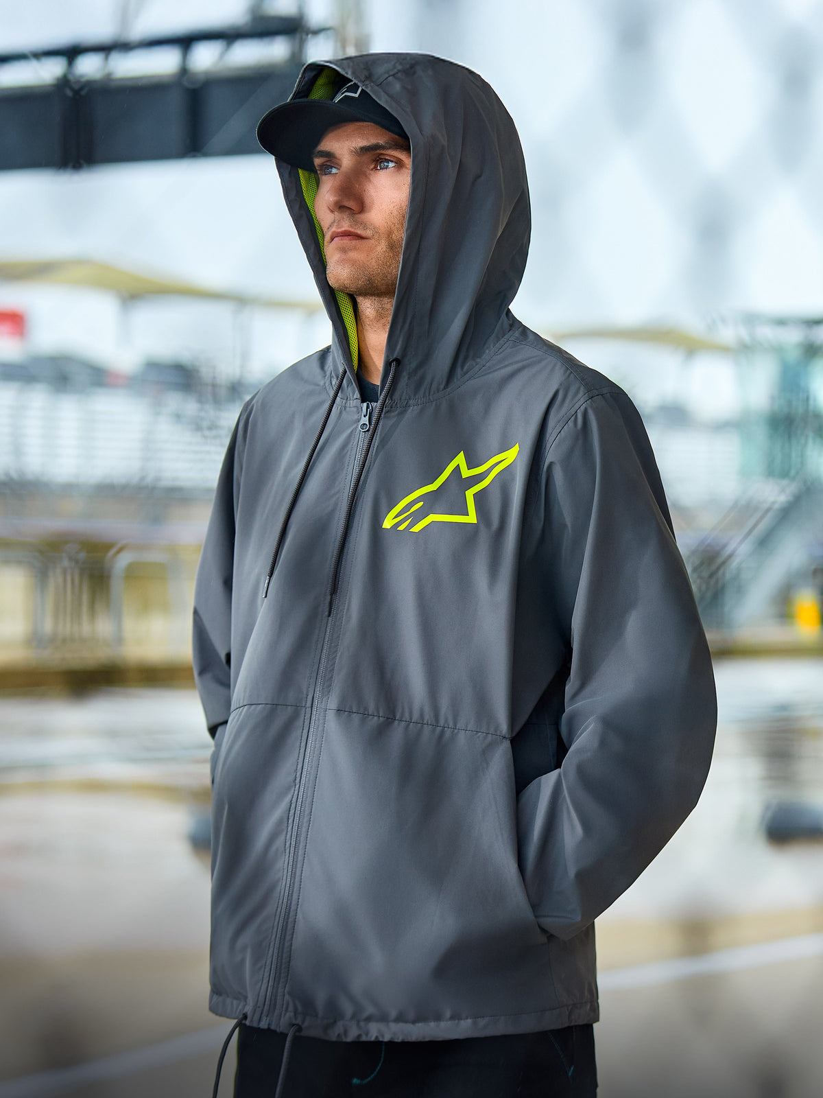 Giacca frangivento Alpinestars Speeder Chromium, giacca lifestyle grigio antracite con accenti giallo alta visibilità, design con cappuccio con fodera interna gialla e logo sul petto, abbigliamento streetwear casual da motorsport indossato da un modello all'aperto