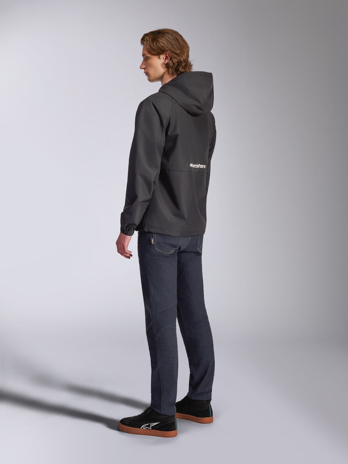 Giacca a vento Alpinestars Speeder Chromium, nera con logo bianco sul retro, giacca casual da uomo con cappuccio, vista di profilo indossata con jeans scuri e sneakers nere, streetwear ispirato al motorsport