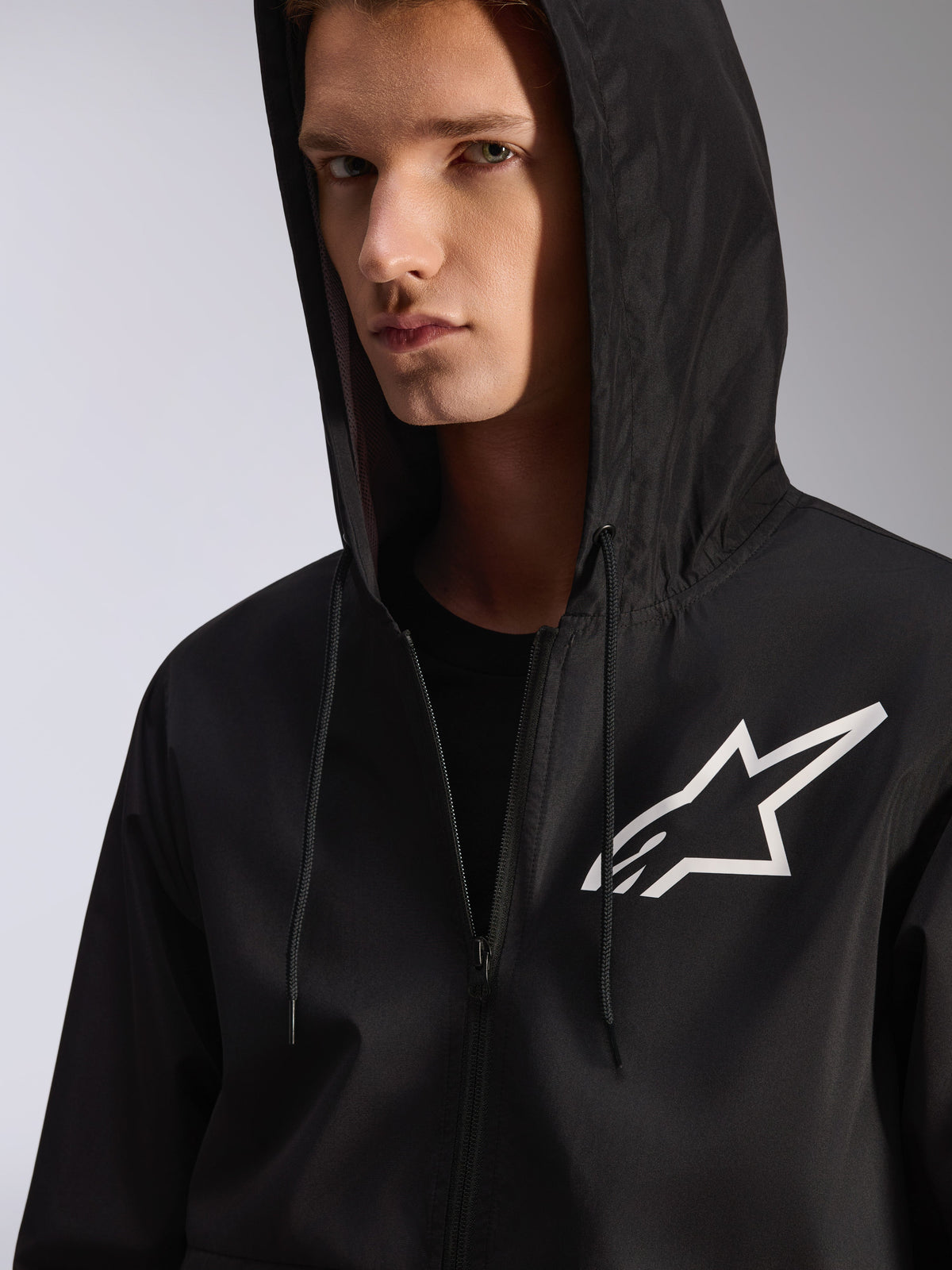 Alpinestars Speeder Chromium Windbreak Jacket, nero, primo piano di una giacca frangivento con cappuccio e logo a stella bianco sul petto, cappuccio con coulisse, materiale tessile leggero, streetwear casual ispirato al motorsport