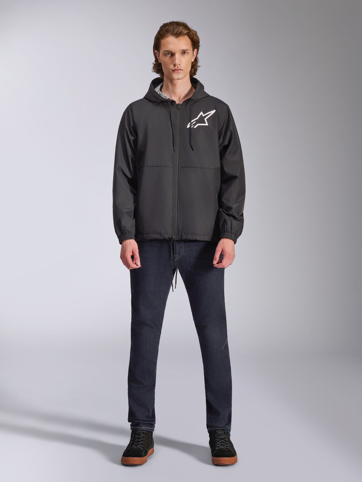 Giacca a vento Alpinestars Speeder Chromium, nera con logo Astars bianco, indossata da un modello con jeans blu scuro e sneakers nere, caratterizzata da un design con cappuccio e cerniera frontale, streetwear casual ispirato al motorsport
