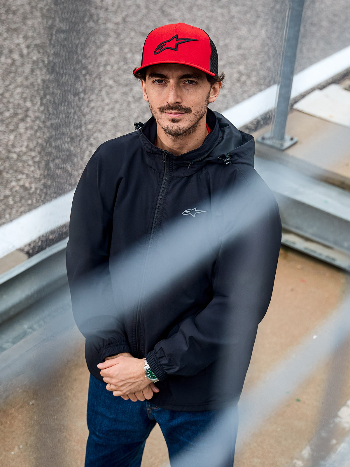 Uomo che indossa la giacca Alpinestars Overhaul Racer in nero, una giacca guscio antipioggia con cappuccio resistente all'acqua e piccolo logo sul petto, abbinata a un cappellino Alpinestars rosso, abbigliamento lifestyle casual motorsport