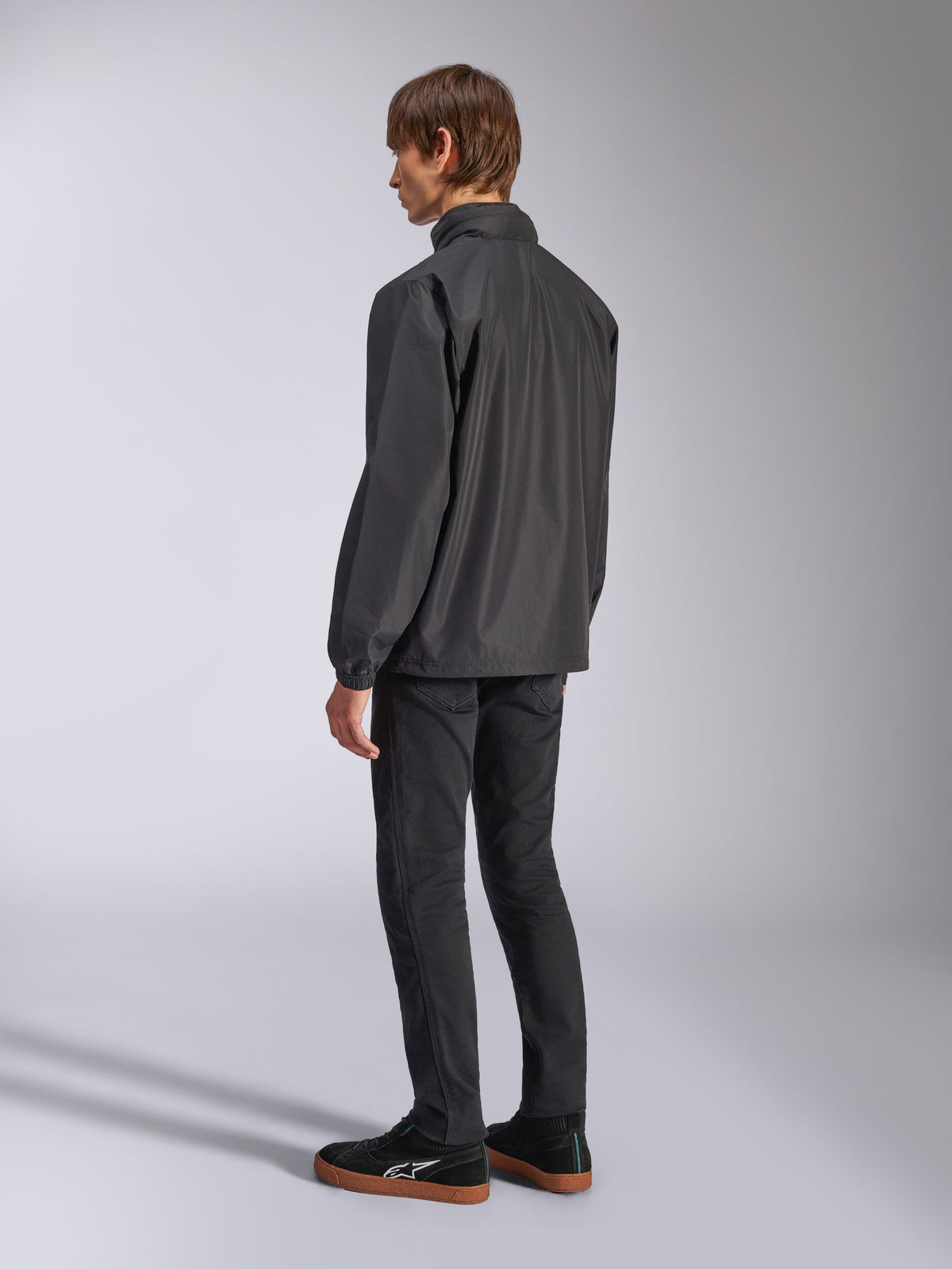 Alpinestars Overhaul Racer Jacket, nera, vista posteriore a tre quarti, indossata da un modello con jeans neri e sneakers nere, giacca guscio antipioggia leggera e resistente all'acqua per uno stile di vita motorsport casual