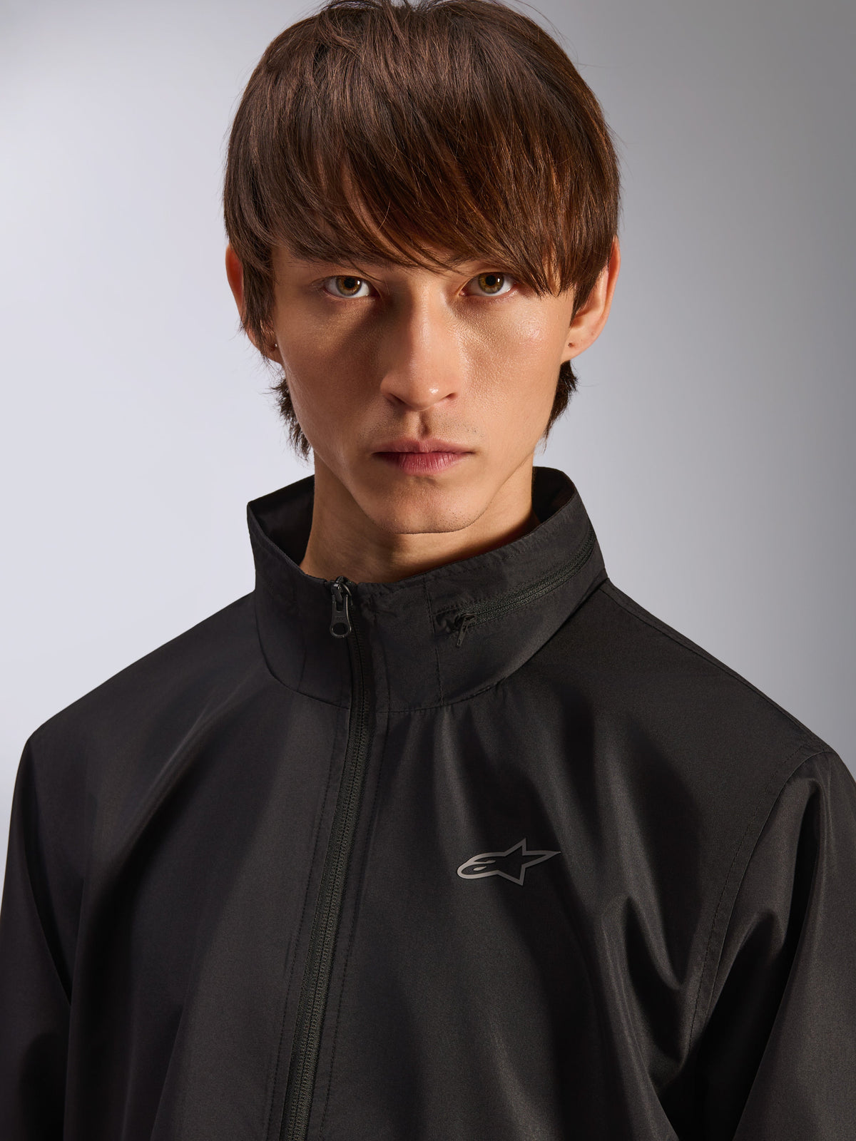 Alpinestars Overhaul Racer Jacket, giacca guscio antipioggia nera, ritratto ravvicinato di un modello che indossa la giacca, con colletto alto e piccolo logo Astars argento sul petto, abbigliamento casual ispirato al motorsport