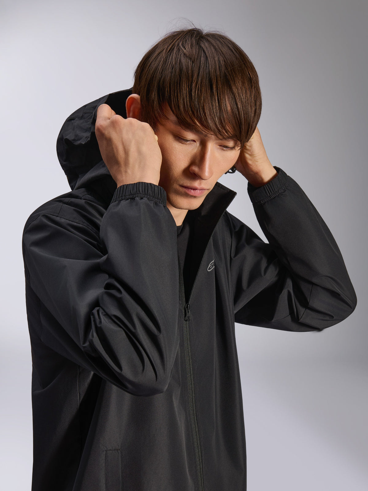 Giacca Alpinestars Overhaul Racer, guscio antipioggia nero con cappuccio, indossata da un modello con cappuccio alzato, realizzata in tessuto 2.5L resistente all'acqua, polsini elasticizzati e logo minimal sul petto, capospalla casual di ispirazione motorsport