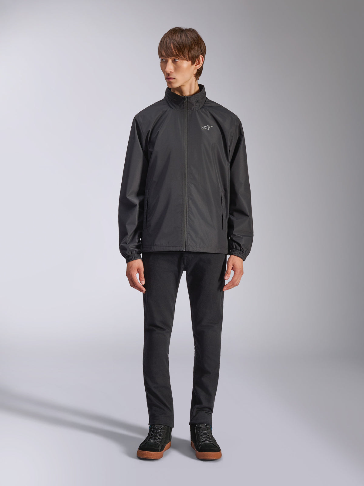 Giacca Alpinestars Overhaul Racer, guscio antipioggia nero da uomo, vista a figura intera su un modello abbinata a pantaloni neri e sneakers casual, con un piccolo logo bianco sul petto, tessuto resistente all'acqua e design casual ispirato al motorsport
