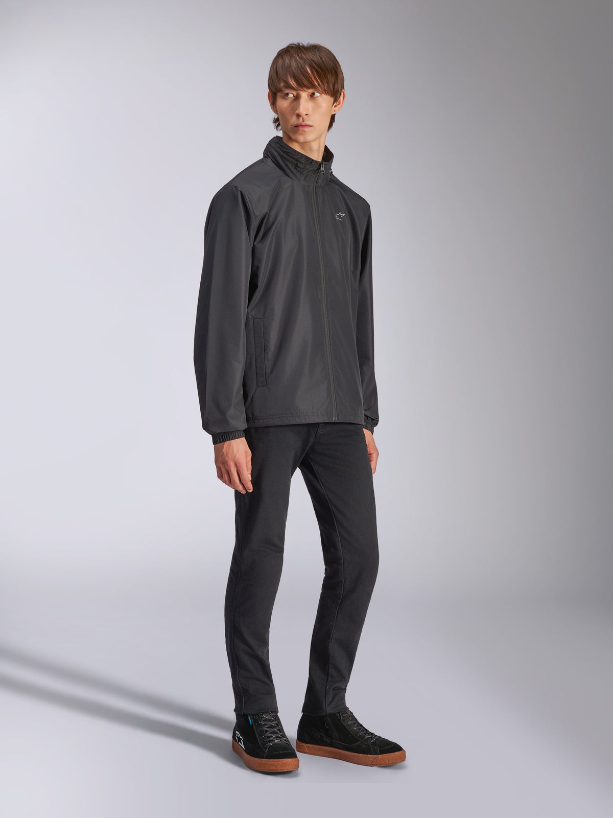 Giacca Alpinestars Overhaul Racer, giacca guscio antipioggia nera, indossata con pantaloni neri e sneakers nere con suola color gomma, con logo a stella bianco sul petto, chiusura frontale con zip, abbigliamento sportivo casual ispirato al motorsport