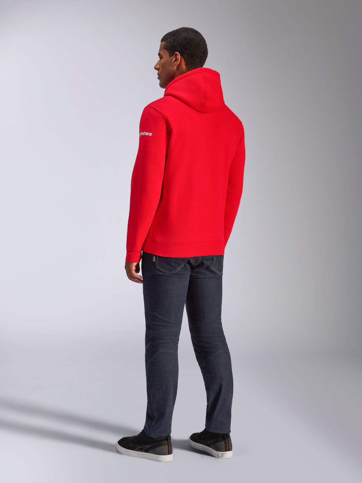 Felpa con cappuccio Alpinestars Frontal, felpa pullover in pile rossa, vista posteriore con logo testuale bianco sulla manica, indossata con pantaloni in denim scuro e scarpe nere, streetwear casual motorsport