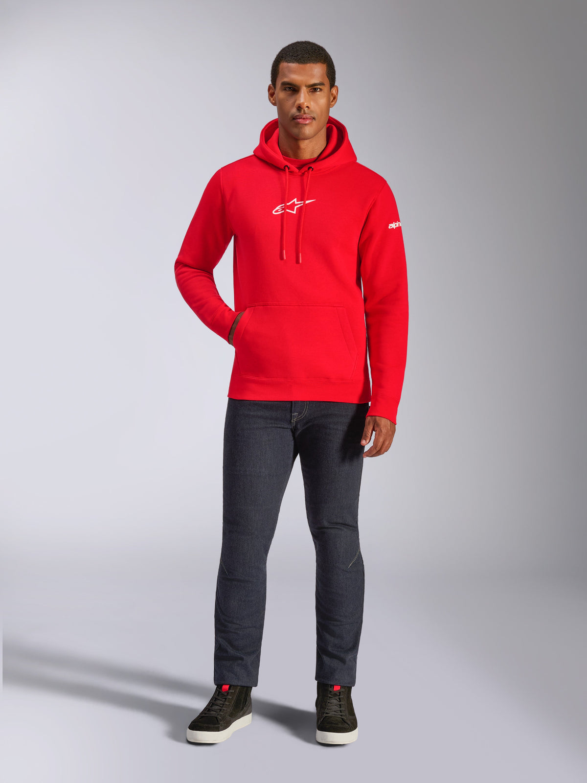 Modello uomo con felpa Alpinestars Frontal Hoodie rossa, felpa pullover in pile con logo Astars bianco sul petto, tasca a marsupio, cappuccio con cordino, abbinata a jeans grigio scuro e sneakers alte nere, streetwear casual motorsport