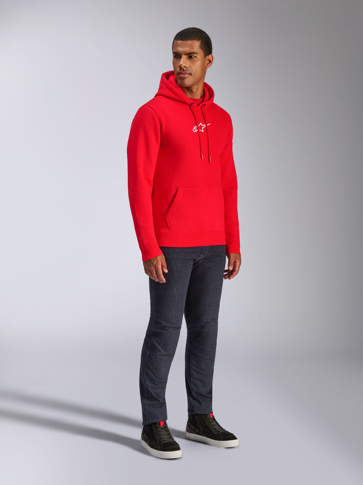 Alpinestars Frontal Hoodie, rossa, modello che indossa una felpa in pile con cappuccio e logo A-star bianco sul petto, tasca a marsupio, cappuccio con cordoncino, abbinata a jeans in denim scuro, streetwear casual ispirato al motorsport