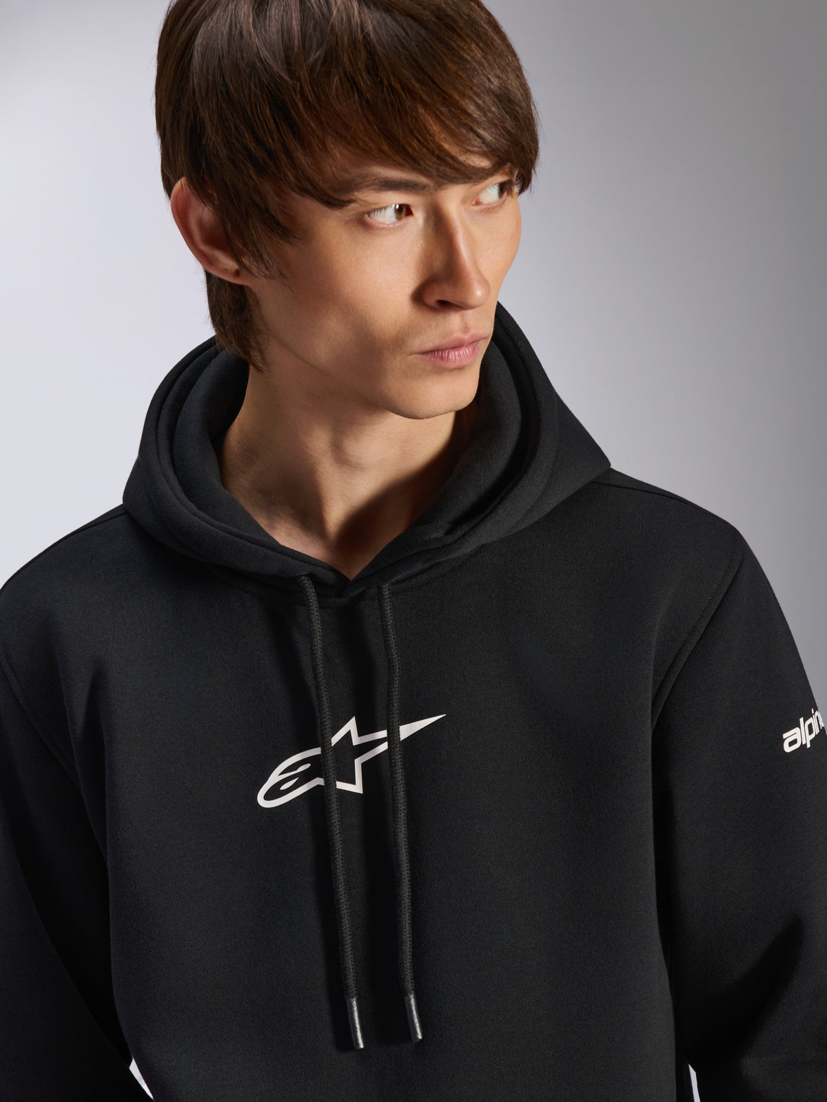 Alpinestars Frontal Hoodie, felpa con cappuccio nera in pile, logo Astar bianco sul petto e branding sulla manica, cappuccio con cordino, indossata da un modello, abbigliamento streetwear casual motorsport