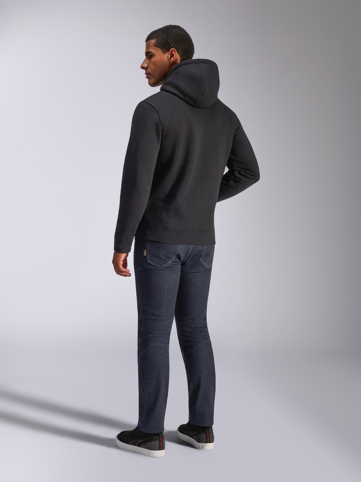 Felpa con cappuccio Alpinestars Blaze V3, vista posteriore di un uomo che indossa la felpa nera, abbinata a jeans scuri e sneakers nere, streetwear casual ispirato al motorsport
