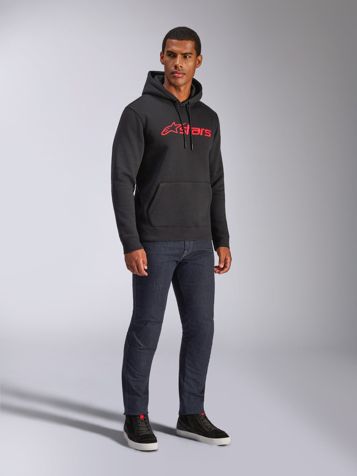 Alpinestars Blaze V3 Hoodie, felpa nera con cappuccio con logo Alpinestars rosso sul petto, indossata da un modello con jeans in denim scuro e sneakers nere, streetwear casual ispirato al motorsport