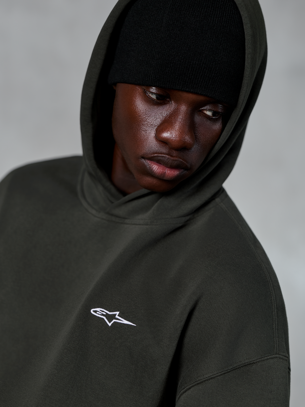 Alpinestars Essentials Ageless Hoodie, Off Black, primo piano di un modello che indossa un berretto nero sotto il cappuccio, con un piccolo logo a stella bianco sul petto, streetwear casual ispirato al motorsport
