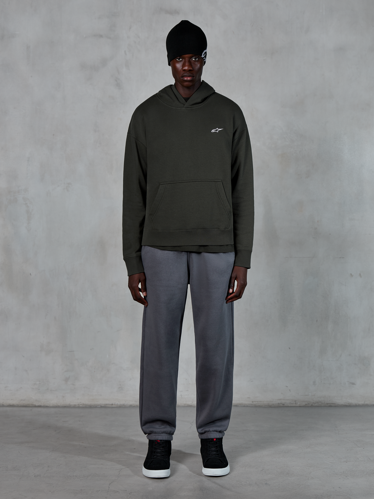 Modello che indossa la felpa con cappuccio Alpinestars Essentials Ageless, streetwear casual, Off Black con piccolo logo bianco sul petto, abbinata a joggers grigi, berretto nero e sneakers nere, design minimalista ispirato al motorsport