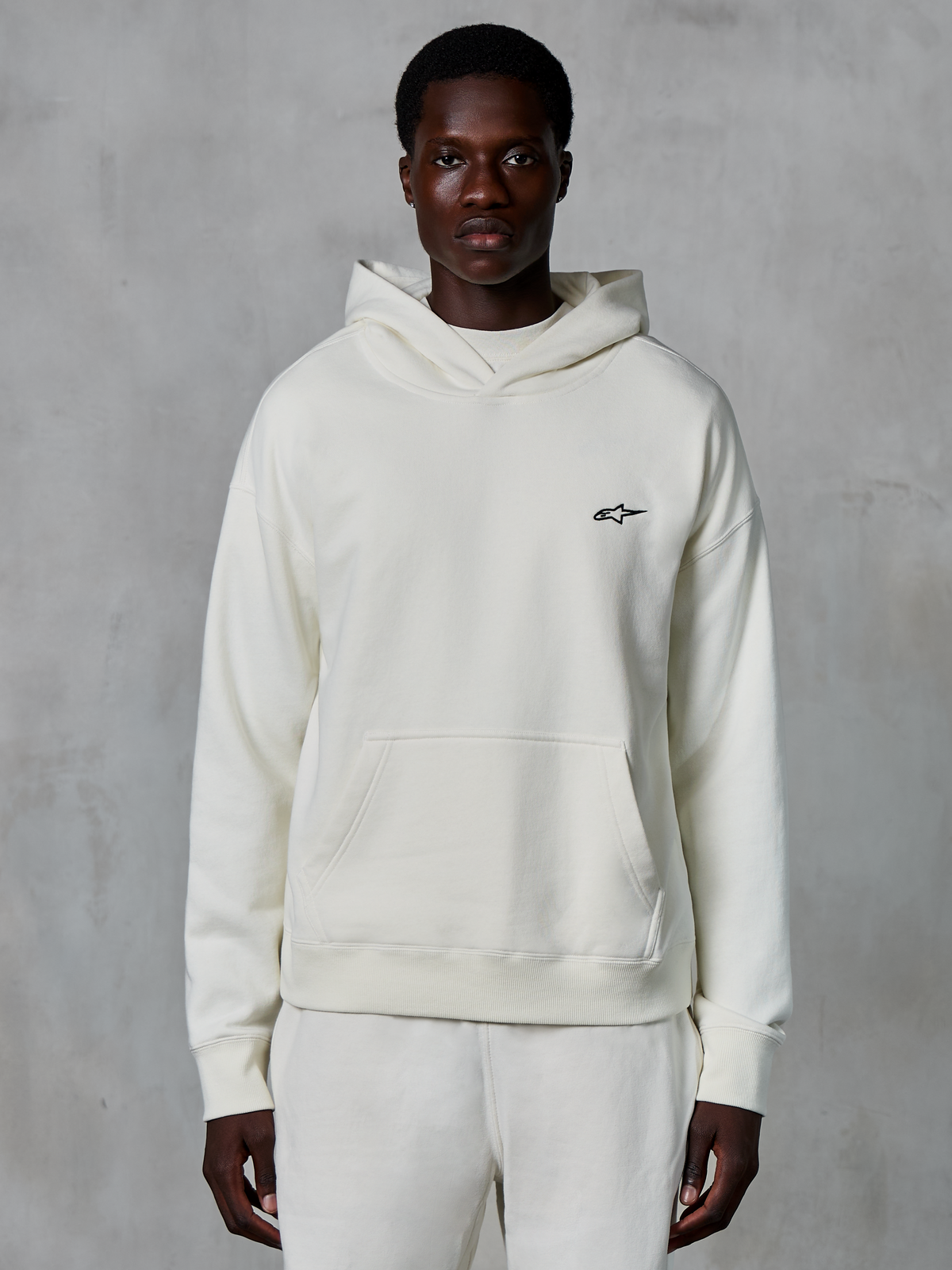 Alpinestars Essentials Ageless Hoodie, bianco sporco, indossata da un modello uomo, piccolo logo nero sul petto, tasca a marsupio frontale, maniche lunghe, streetwear casual ispirato al motorsport