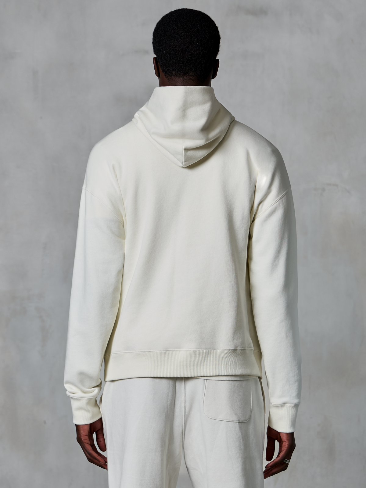 Alpinestars Essentials Ageless Hoodie, felpa con cappuccio pullover off-white, vista posteriore su modello, design pulito e minimalista, pantaloni casual coordinati, vestibilità rilassata per il comfort quotidiano